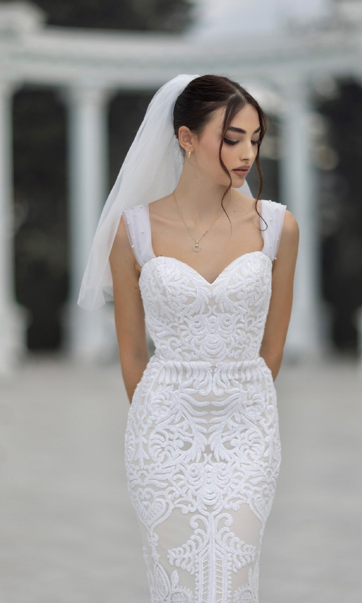 A bride wearing the Off-Shoulder White Bridal Gown: Tina Holly TW103 with lace and veil stands outdoors, looking down with her hands behind her back. Blurred marble columns are visible in the background.